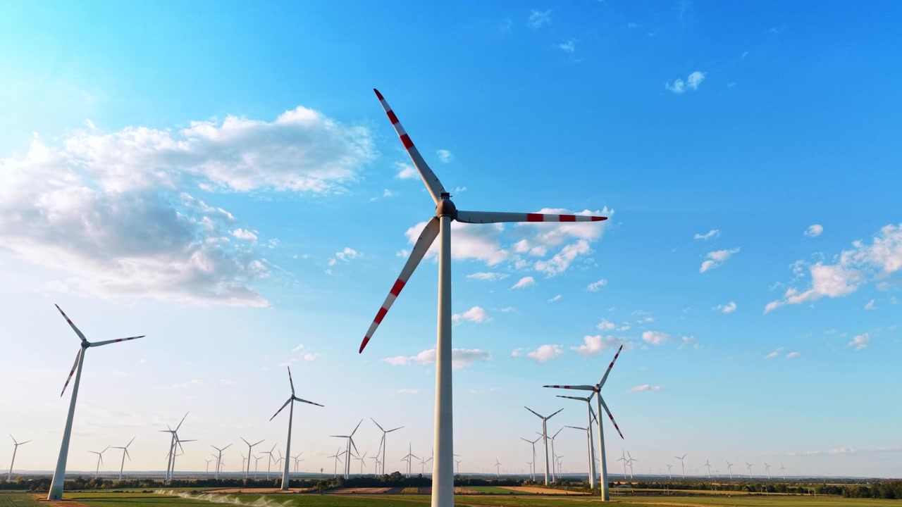 Large wind turbine towering into a clear blue sky. A low angle shot shows the steel tower and red striped blades of a modern wind generator over a distant horizon.