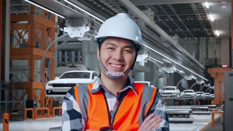 primer plano de un ingeniero asiático con casco de seguridad cruzando los brazos y sonriendo a la cámara mientras está de pie en una fábrica de automóviles ev