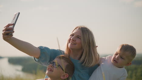 a woman in a blue dress sits on the grass, as she takes a picture with her smartphone. her two sons, both wearing white shirts, are with her; the younger son appears unhappy while the older one smiles