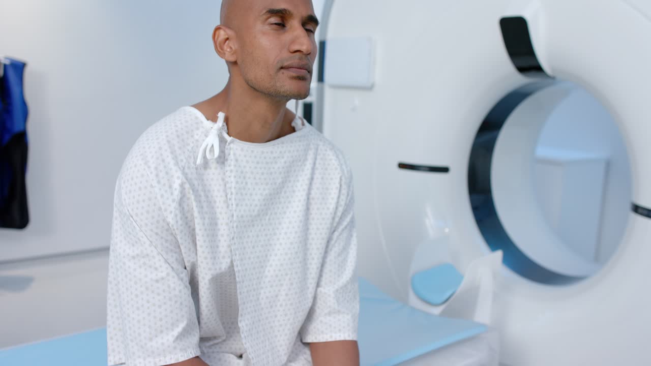 Uomo africano che stringe e sfrega le mani seduto sul lettino radiologico, indossa un camice ed è in preparazione per una TAC