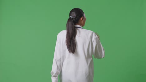vista trasera de una doctora asiática con un estetoscopio agitando la mano y sonriendo mientras camina en el fondo de la pantalla verde en el hospital