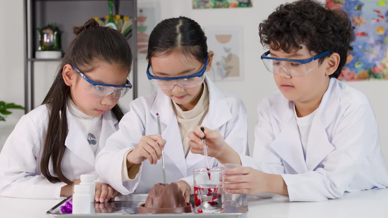 Tiga anak Asia mengenakan jas lab dan kacamata pengaman melakukan percobaan kimia di dalam kelas.