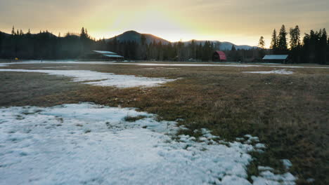 Aerial-fast-paced-flying-low-over-snowy-farmland-field-at-sunset,-rising-up-over-ranch-barns-and-cabins