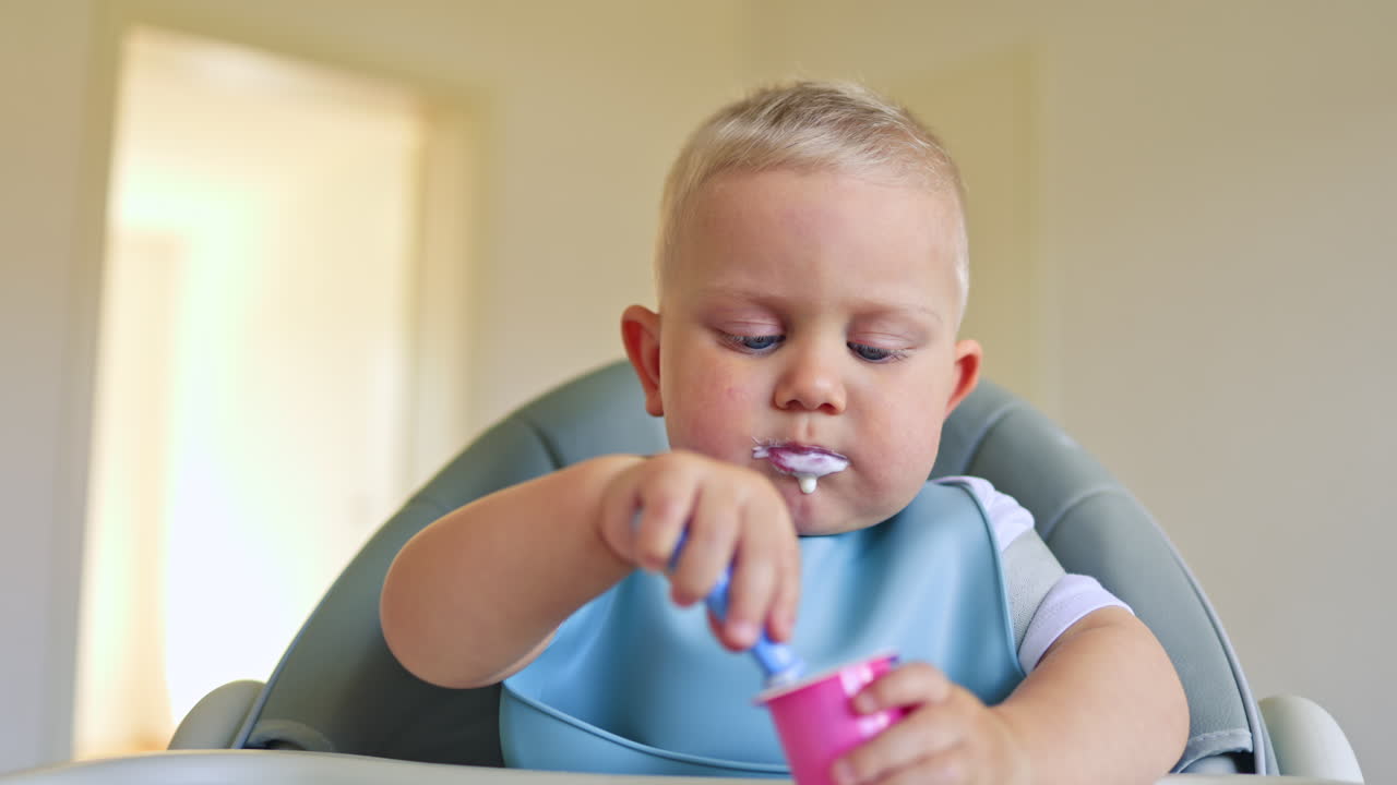 Two-year-old boy sits and eats yogurt independently. Toddler boy learns to eat independently. Dairy products are waiting for children