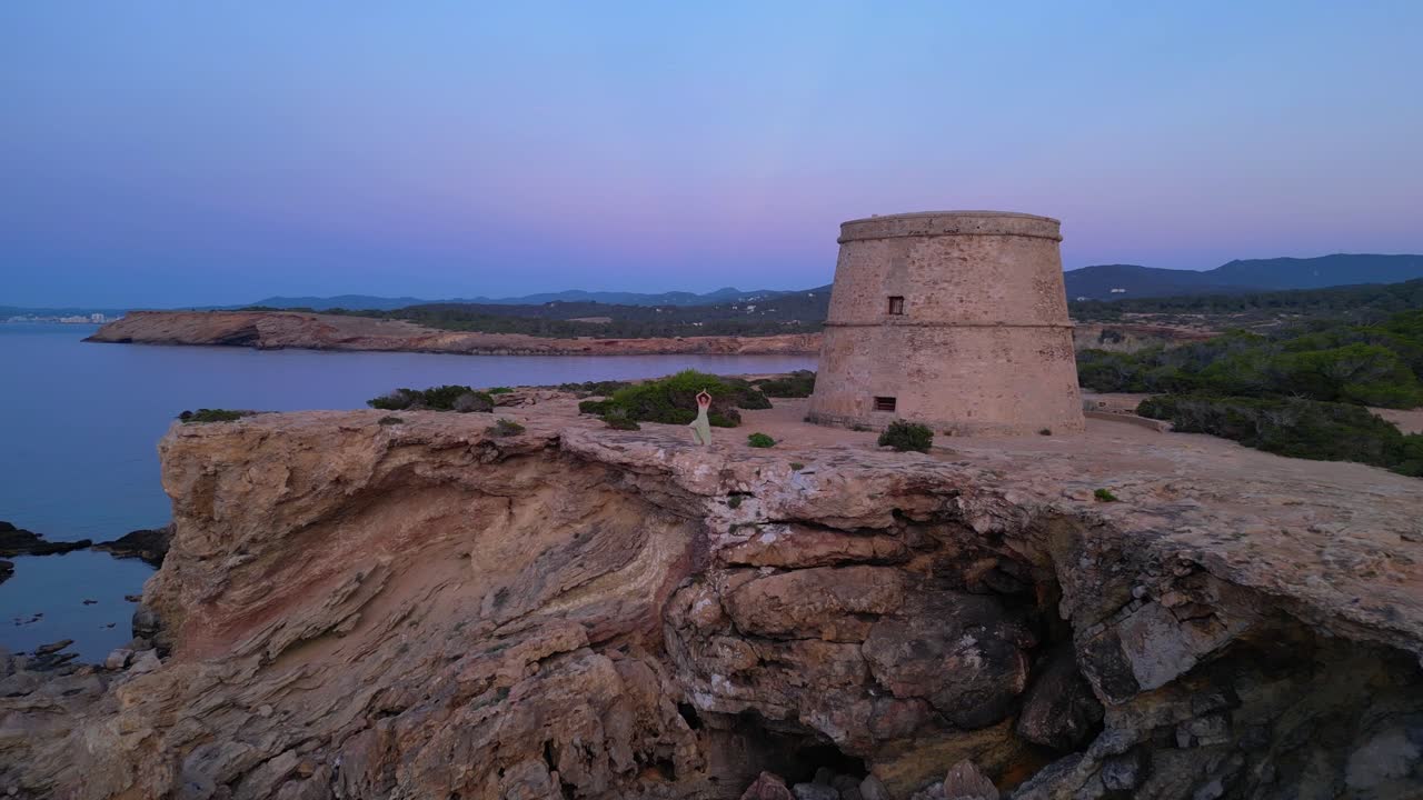 Barefoot hippie girl practicing yoga at sunset near Torre des Carregador, Ibiza, Spain. Stunning aerial view flight pull in drone