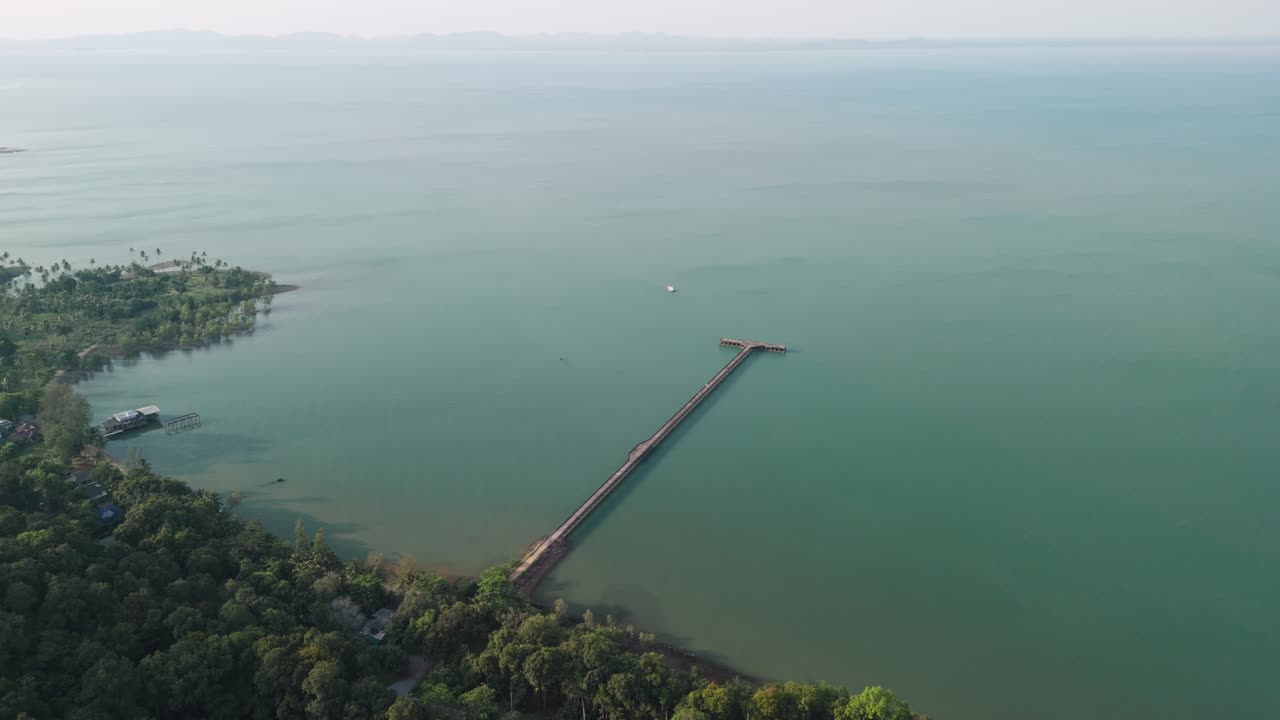 Drone descending toward wooden jetty extending into turquoise bay surrounded by dense jungle in misty tropical conditions