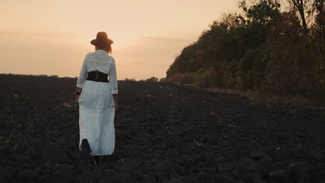 a woman in a dress walks through a plowed field on black soil