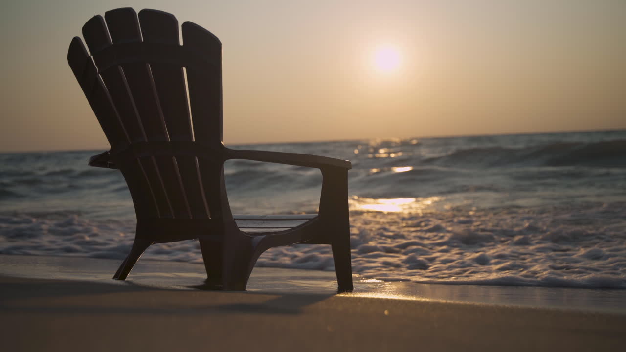 Silhouette of an empty Muskoka chair sits on a beach as the sun sets in the background