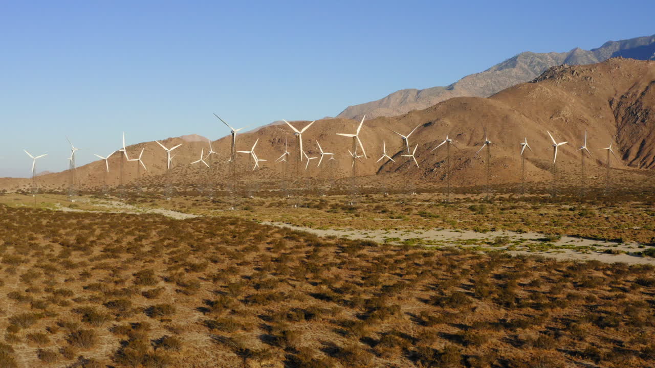 drone volando hacia docenas de turbinas eólicas en un enorme parque eólico con montañas en el fondo cerca de palm springs en el desierto de mojave, california, ee.uu.
