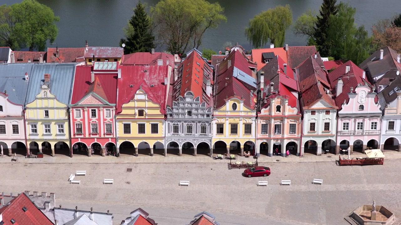 Vista aérea de drone de uma fileira de casas geminadas renascentistas coloridas com arcadas ao longo da praça principal de uma cidade checa histórica com um lago visível ao fundo, Telč, República Checa