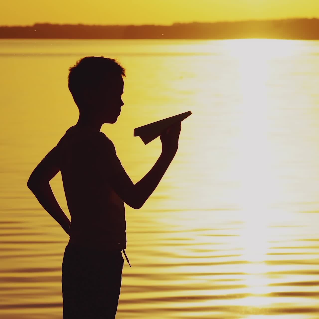 A little boy is holding a paper airplane in his hand and playing with him on the bank of the river at sunset on the background of the solar track.