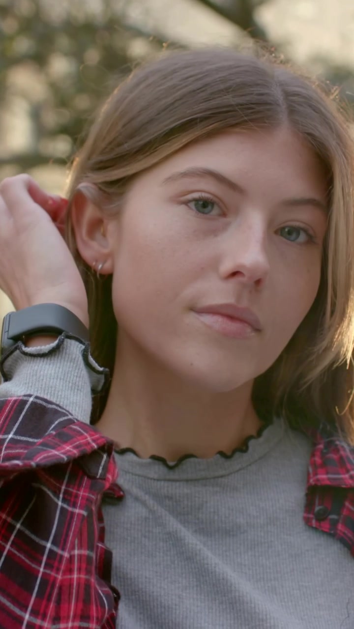 Video verticale: Donna che si gira dopo il segnale della telecamera, si sistema i capelli al parco, con camicia a quadri e smartwatch in vista.