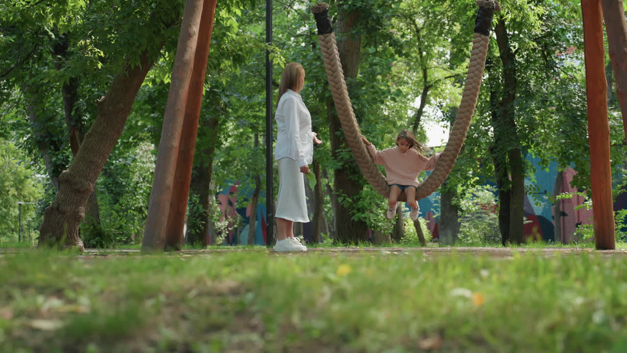 Mamá observa a su hija balancearse tranquilamente bajo los árboles, con un dosel verde suave, la madre de pie cerca con ropa blanca, la niña relajada en una hamaca de cuerda, brisa suave y un momento familiar tranquilo