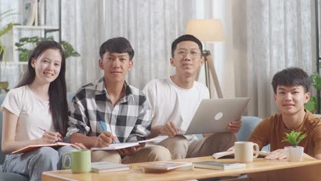 asian teen group smiling to camera while studying at home, doing project, a girl and a boy in plaid shirt writing into notebooks, a boy in white t-shirt typing on laptop sitting next to another boy reading