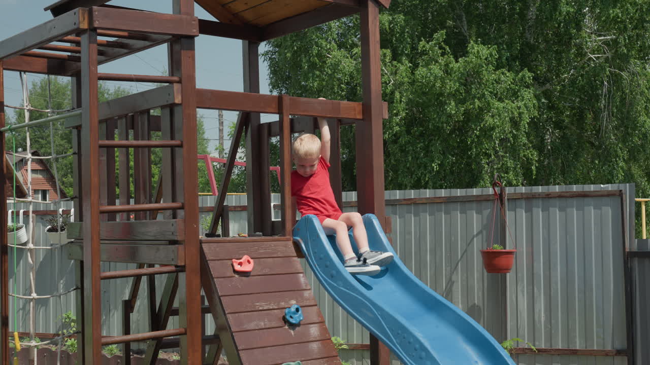Young Child Slides Down Colorful Backyard Slide, Boy With Blond Hair Has Fun On Garden Playground Equipment, Happy Young Boy With Light Hair Descends Brightly Colored Slide In Backyard Garden Area