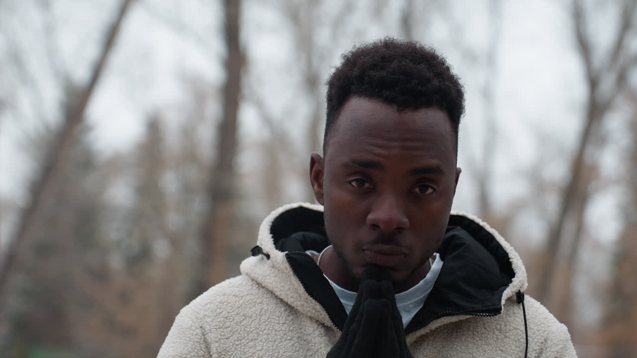 Young boy in black gloves and white winter jacket standing outdoors during winter, hands pressed together in deep thought, with a blurred background of bare trees and snow