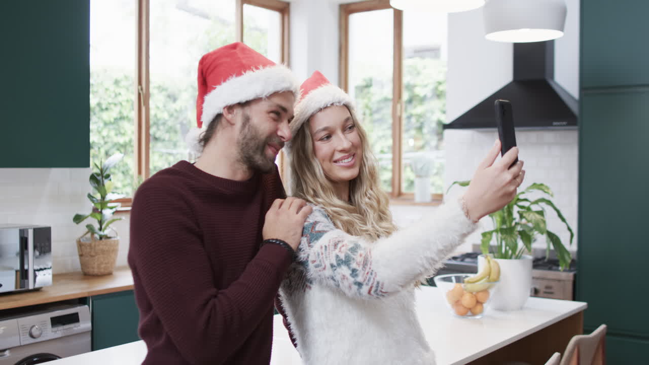 una pareja diversa con sombreros de santa usando un teléfono inteligente y tomando selfies, en cámara lenta