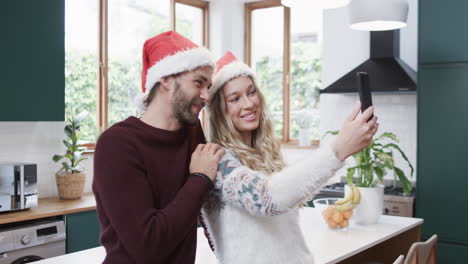 Pareja-Diversa-Usando-Sombreros-De-Santa-Usando-Un-Teléfono-Inteligente-Y-Tomando-Selfies,-En-Cámara-Lenta