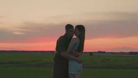 young couple stands face to face holding hands in peaceful field during glowing sunset, gazing into each other eyes with warm expressions while surrounded
