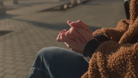 close-up of a person sitting with face not visible, wearing blue jeans and a brown coat. the hands are tightly clasped together in a gesture of anxiety or deep thought, conveying tension or worry