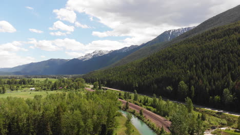 aerial shot of road, river, and train tracks winding through east glacier park, montana, snow-capped mountains and forested hills in the background