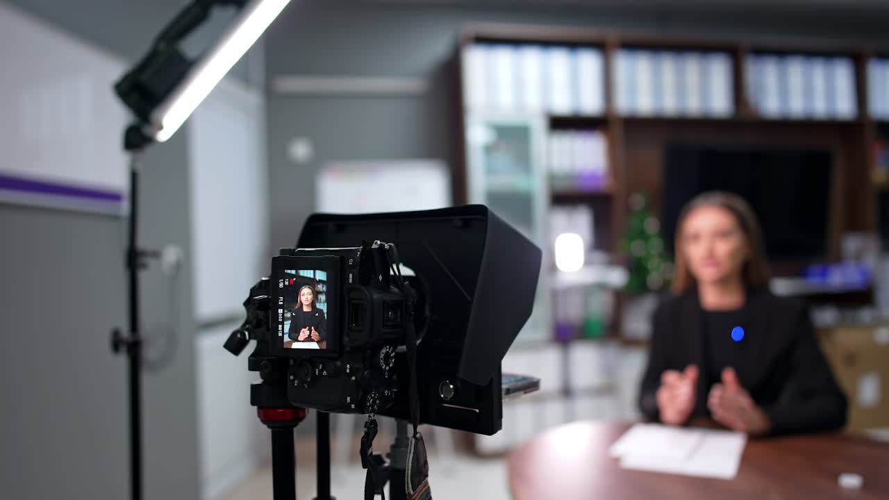 Confident reserved Caucasian female in black jacket sits at desk talking. Woman records video for blog on professional camera. Blurred backdrop.