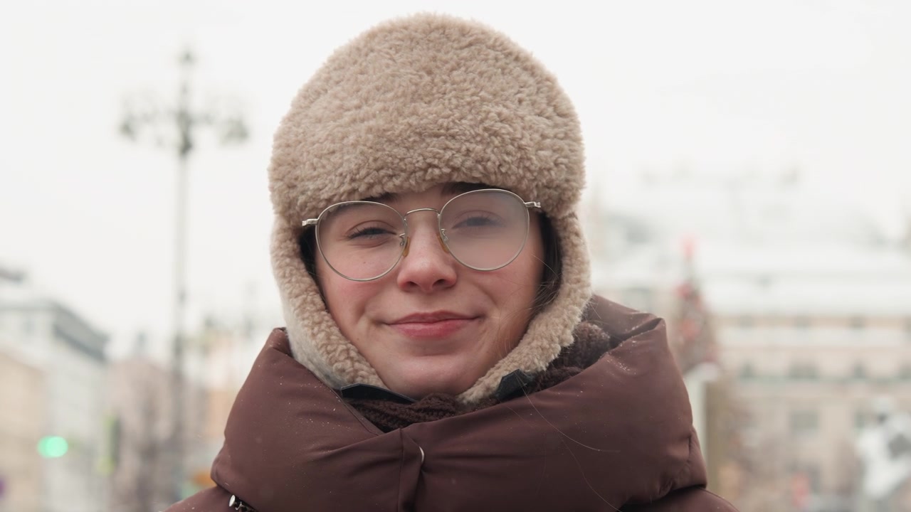 Close-up head-and-shoulders view of young person outdoors on chilly city street, bringing hands to mouth to warm fingers, then relaxing into gentle smile with blurred urban background and soft