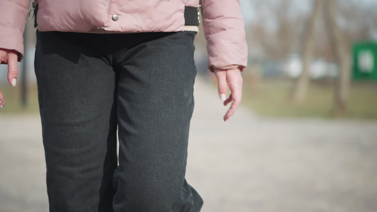 Close-up frame shows young woman walking outdoors in cold weather, with focus on left arm, pink winter sleeve, relaxed hand, and black denim pants as she steps forward along blurred paved park path