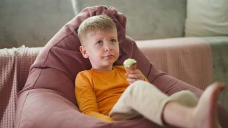 young boy lounges comfortably on large pink beanbag chair, wearing orange shirt and light pants, holding ice cream cone with chocolate and mint, gazing thoughtfully upward