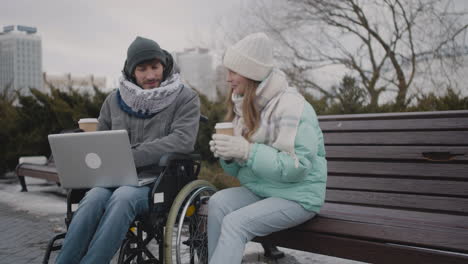 Behinderter-Mann-Im-Rollstuhl-Und-Sein-Freund-Sehen-Sich-Etwas-Lustiges-Auf-Dem-Laptop-An,-Während-Sie-Im-Winter-1-Im-Stadtpark-Kaffee-Zum-Mitnehmen-Trinken