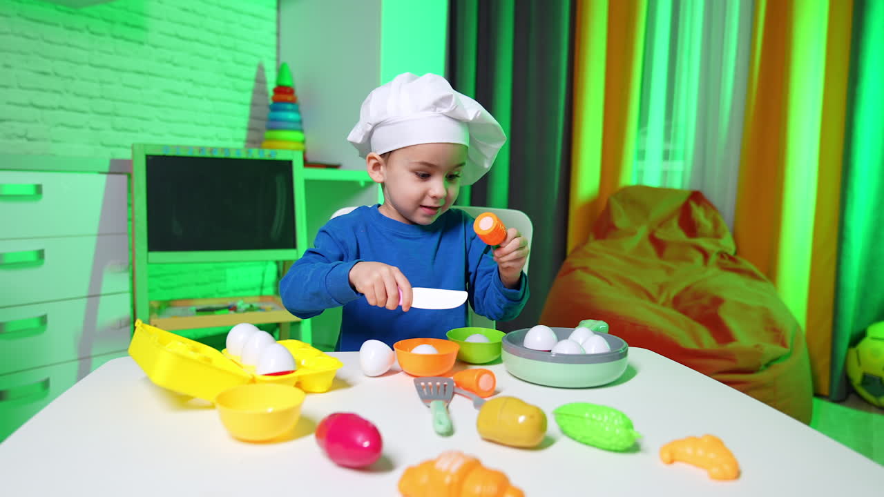 Small baby boy in white cap sits at desk. Smiling toddler plays with toy vegetables.