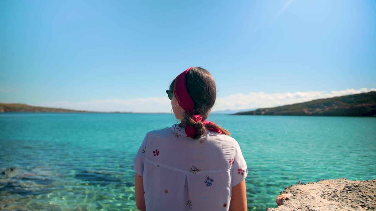 Brunette with fuschia headband looking at transparent turquoise ocean, scratching knee, close shot.