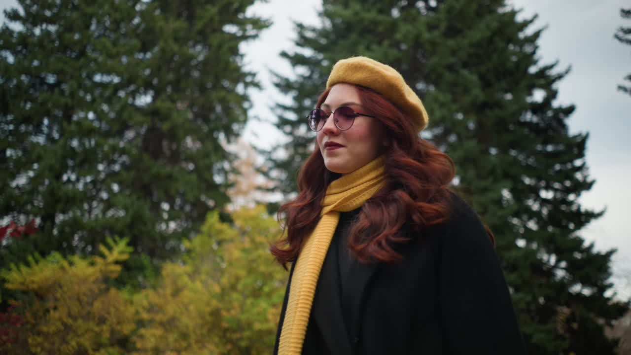 молодая женщина с длинными волнистыми красными волосами, одетая в черное пальто и желтый берет с соответствующим шарфом, настраивает очки, исследуя яркий парк, наполненный красочной осенней лиственнией.