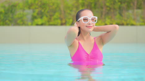 Young-brunette-asian-woman-standing-in-the-swimming-pool-wearing-a-purple-bikini,-touching-her-wet-hair-rising-her-face-up-with-delight,-and-opening-her-eyes,-slow-motion