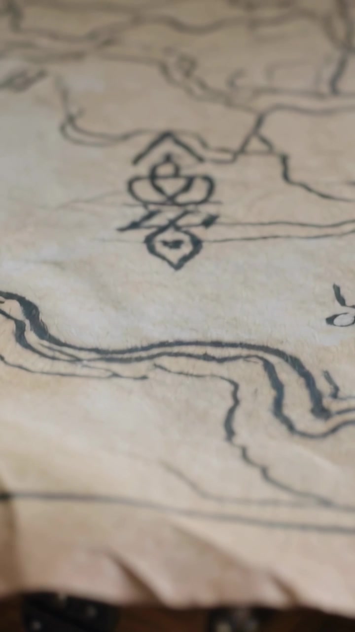 Video verticale: La telecamera si solleva rivelando monete d'oro e un forziere su una scrivania scura, mettendo a fuoco un simbolo ornamentale su una mappa.