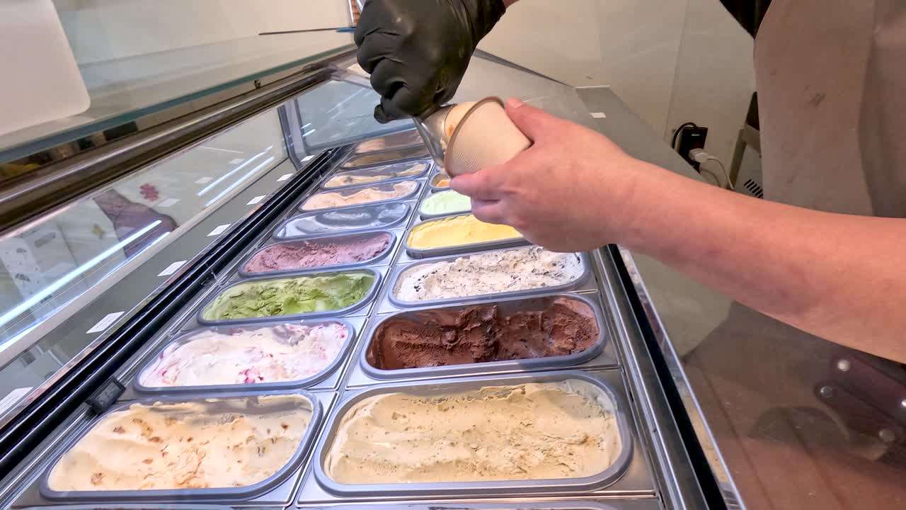 Gloved server scoops colorful gelato into cups at modern dessert shop under bright indoor lighting