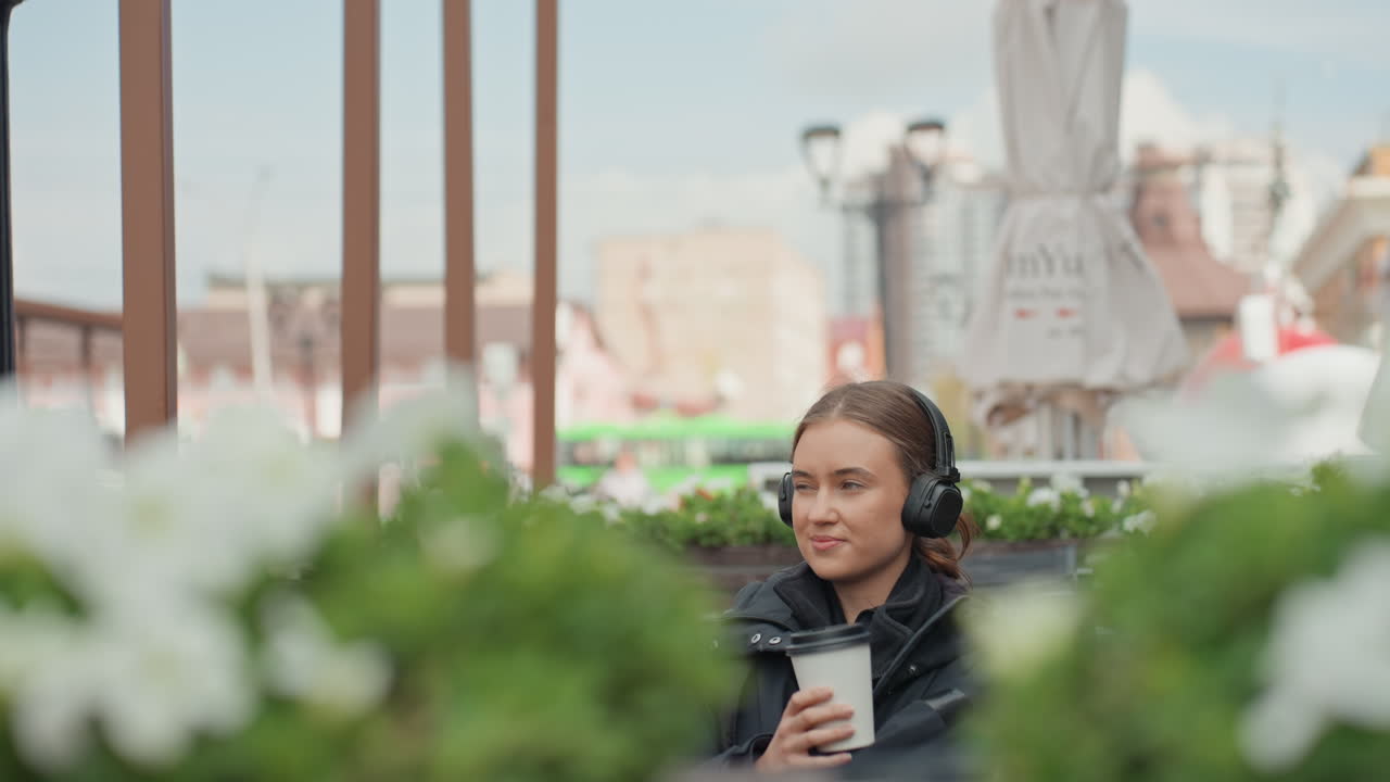 rooftop cafe caucasian woman with headphones wandering among planters and sipping coffee, calm pensive expression, casual jacket, overcast urban skyline, candid portrait suggesting student or florist