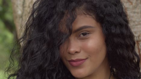 Close-up-of-a-young-woman-with-red-lipstick-smiling-with-a-large-tree-trunk-in-the-background