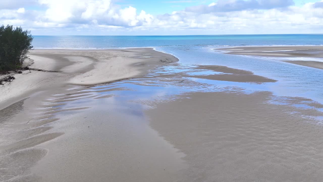 Drone glides above tidal sandbanks, clear water, and rainforest edge under bright daylight