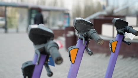 vista cercana de scooters eléctricos púrpuras estacionados en un área ocupada de la ciudad, el fondo incluye detalles borrosos del paisaje urbano con coches en movimiento y personas