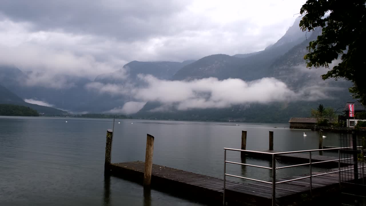 Nuvole basse scivolano sulle montagne, gettando un velo sognante sul lago calmo, mentre un tranquillo pontile di legno si estende nelle serene acque alpine.