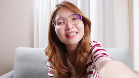 portrait of a selfie asian woman with eyeglasses is smiling with laughter being positive emotions on video call online sitting on the sofa in the living room