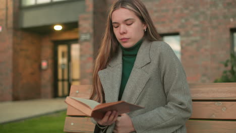 Lady seated on wooden bench adjusting coat while holding open book in hand, calm and reflective expression with modern brick building in background