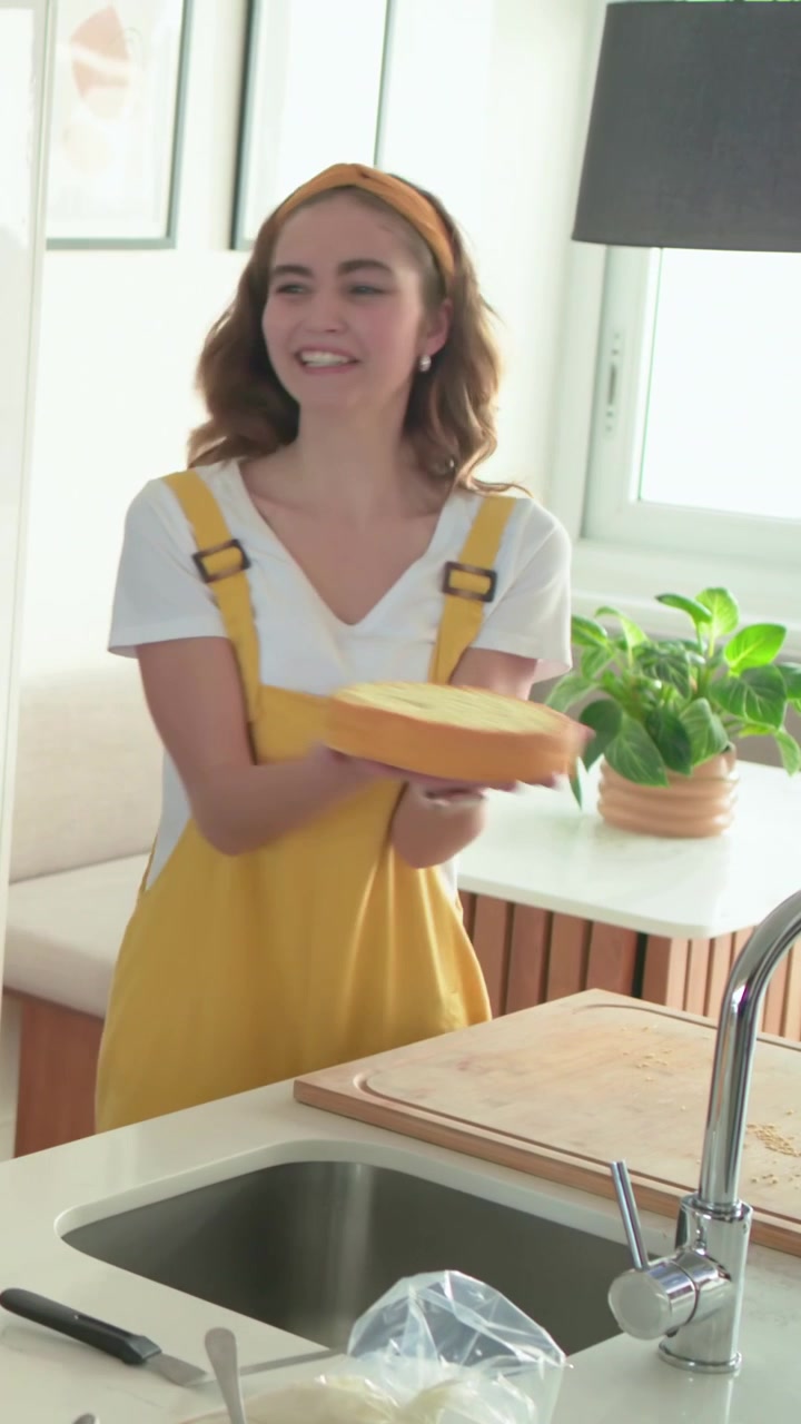 Video verticale: Donna in salopette gialla che raccoglie la glassa e leviga una torta su un'isola della cucina, presentandola.