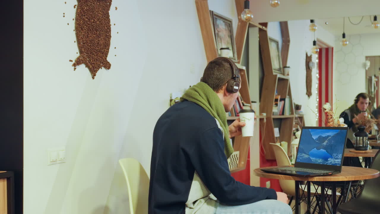 Kaukasische man in gezellig café typt op laptop met koptelefoon, groene sjaal en trui, nipt aan koffie terwijl barista voorbijloopt, warm houten interieur, feestelijke verlichting, ontspannen creatieve werksfeer.