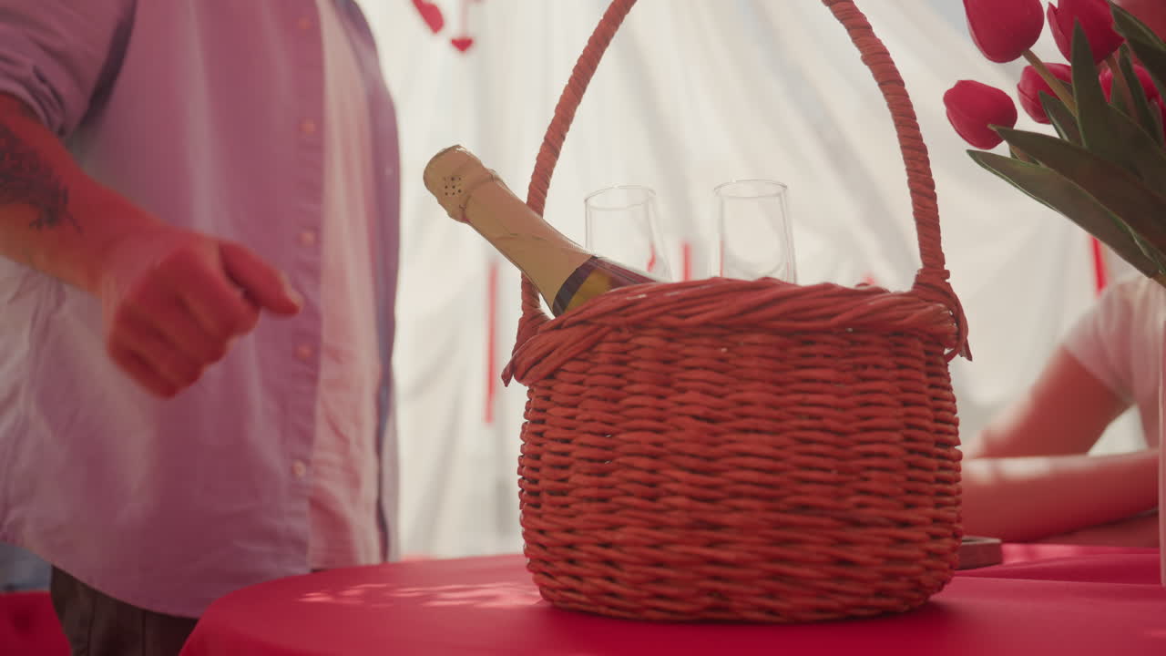 Romantická pikniková scéna: proutěný košík, láhev vína, tulipány na červeném ubrusu uvnitř bílého stanu. Muž umisťuje košík, zatímco partnerka sedí s krajkovým rukávem. Láhev šampaňského vykukuje z košíku vedle dvou sklenic.