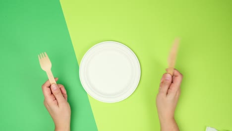 empty white paper plate and female hands are holding disposable fork and knife on a green background