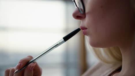 primer plano de una mujer joven girando suavemente la pluma sobre los labios, golpeando suavmente contra los labios en un gesto pensativo, con fondo borroso, capturado en un momento intelectual o creativo