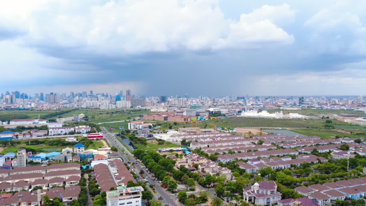 Aerial footage of Phnom Penh's sprawling cityscape under dramatic cloud cover. Captivating bird's eye view, vibrant greens and urban layout under moody skies.