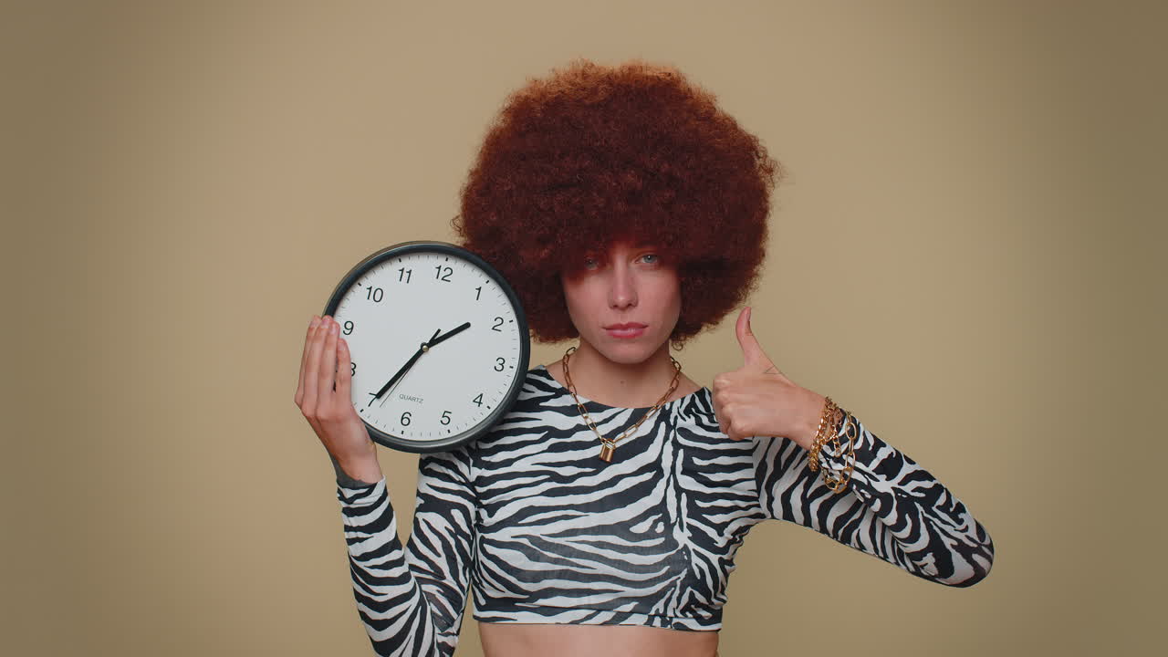 Pretty woman showing time on wall office clock, ok, thumb up, approve, pointing finger at camera